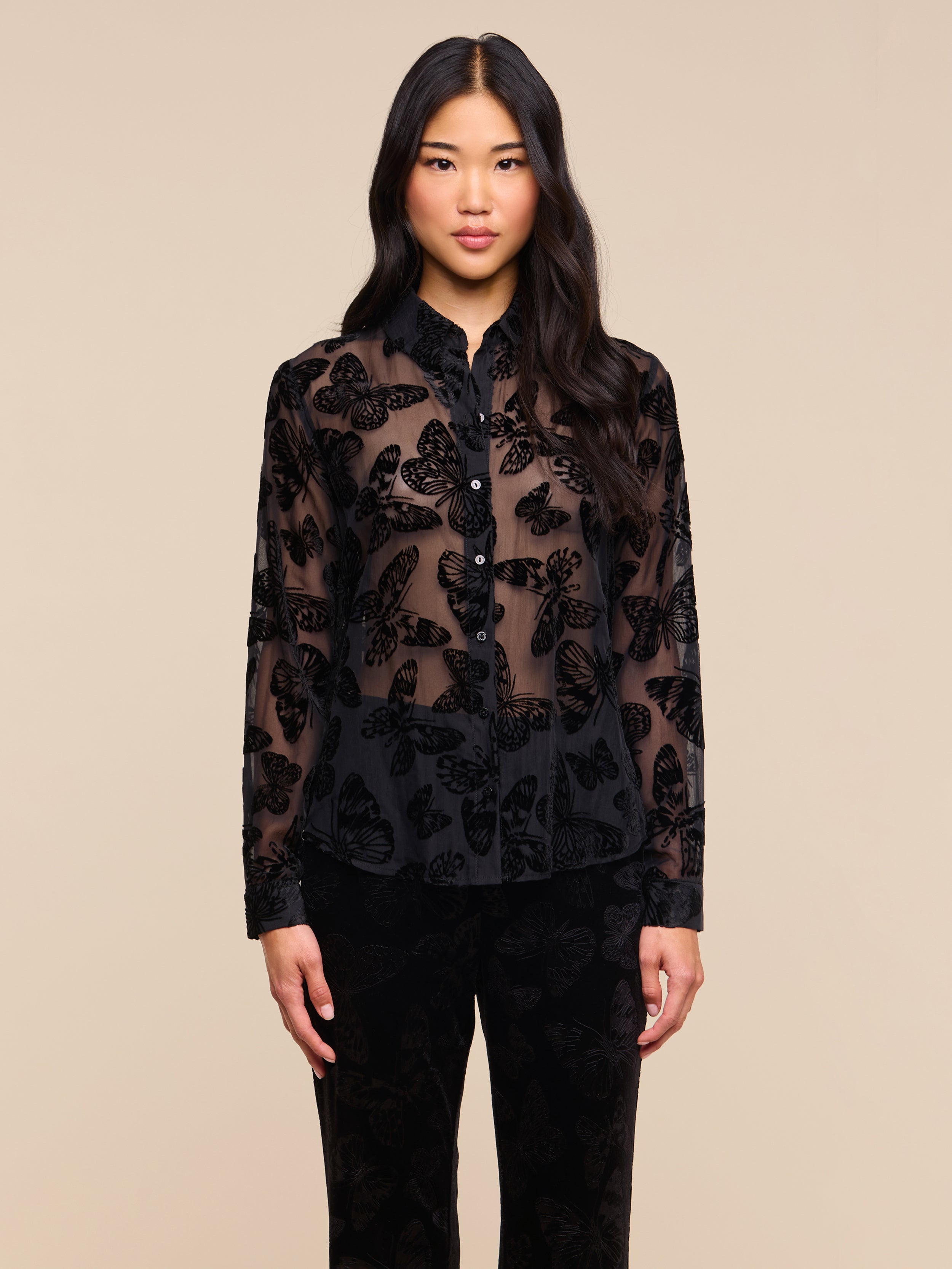 A woman with long black hair stands facing forward against a plain light background, wearing the Laurent Velvet Blouse—featuring a burnout velvet butterfly pattern—paired with black pants.