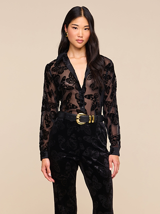 A woman with long hair and a neutral expression stands against a beige background wearing the Laurent Velvet Blouse, a relaxed fit, sheer black top decorated with a delicate butterfly print. She pairs it with black velvet pants and accessorizes with a black belt featuring a large gold buckle.