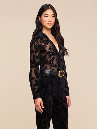 A woman with long black hair wears the Laurent Velvet Blouse—a relaxed fit top crafted from sheer black fabric with velvet butterfly patterns—paired with black pants, a black belt featuring a gold buckle, and gold earrings, standing against a plain beige background.