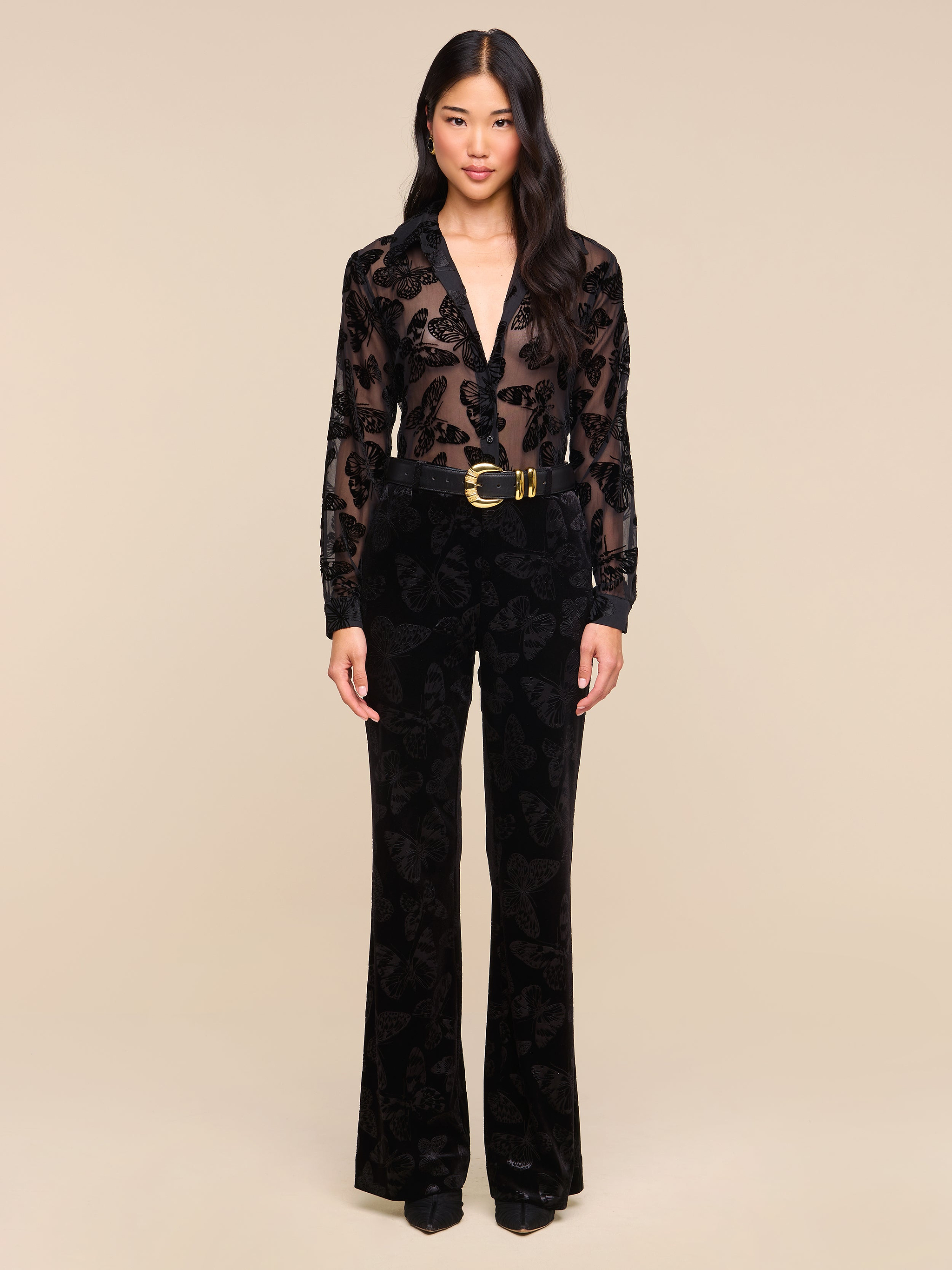 A woman stands facing forward against a plain white background, wearing the Laurent Velvet Blouse—a sheer burnout velvet top with a floral pattern—paired with black pants, a wide black belt featuring a gold buckle, and black shoes.