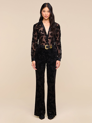 A woman stands against a beige background, wearing the Laurent Velvet Blouse—a sheer black burnout velvet top with a butterfly print—paired with black high-waisted pants featuring a similar pattern and accessorized with a wide black belt with a large gold buckle.