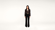 A woman stands facing forward in a black outfit with a floral pattern, featuring a belted jacket and wide-leg pants, against a plain white background.