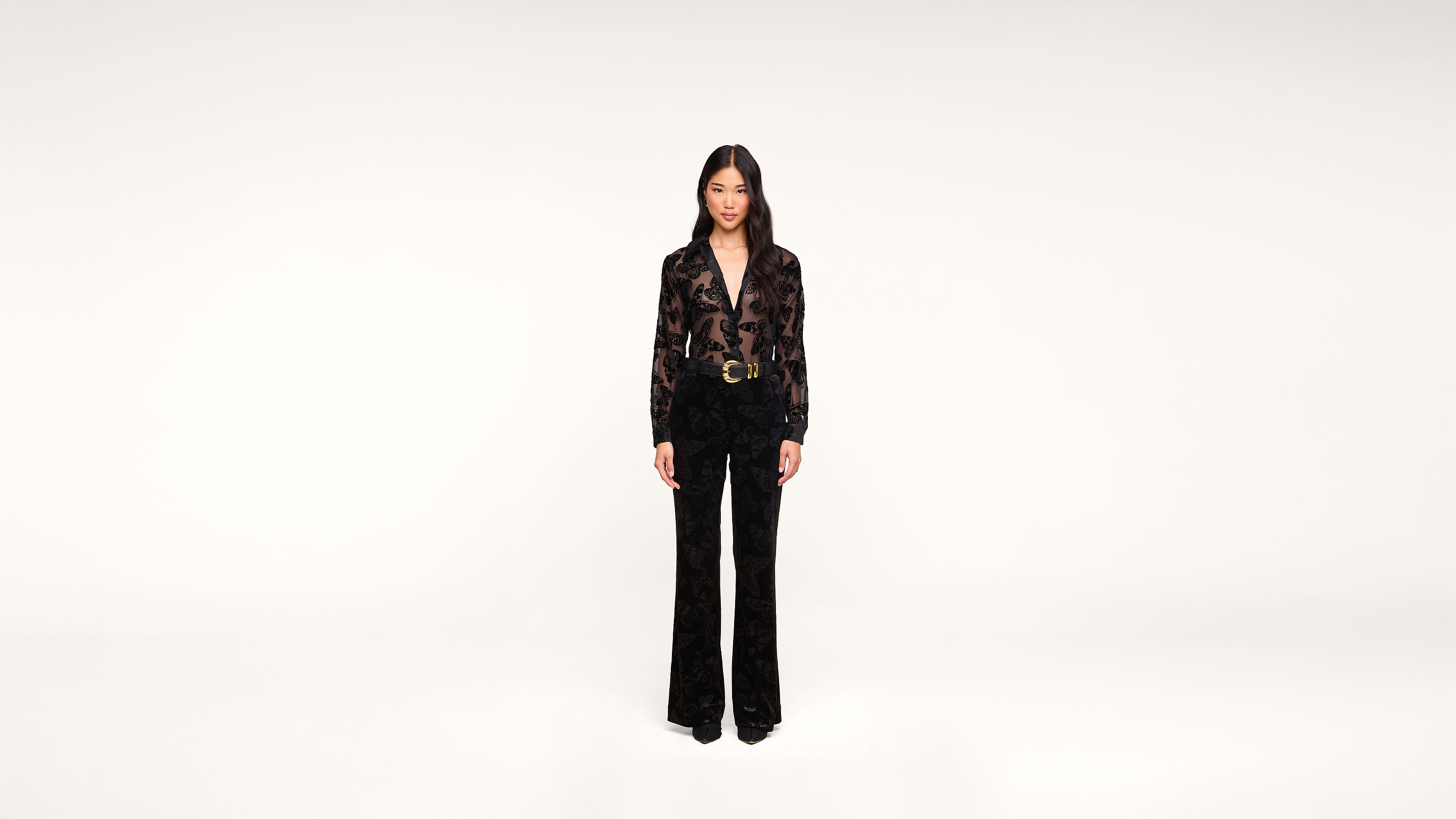A woman stands facing forward in a black outfit with a floral pattern, featuring a belted jacket and wide-leg pants, against a plain white background.