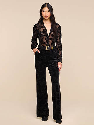 A woman stands against a beige background, wearing the Laurent Velvet Blouse—a sheer black burnout velvet top with a butterfly pattern—paired with black velvet pants, a wide belt featuring a gold buckle, and black shoes. Her long, wavy hair frames her neutral expression.