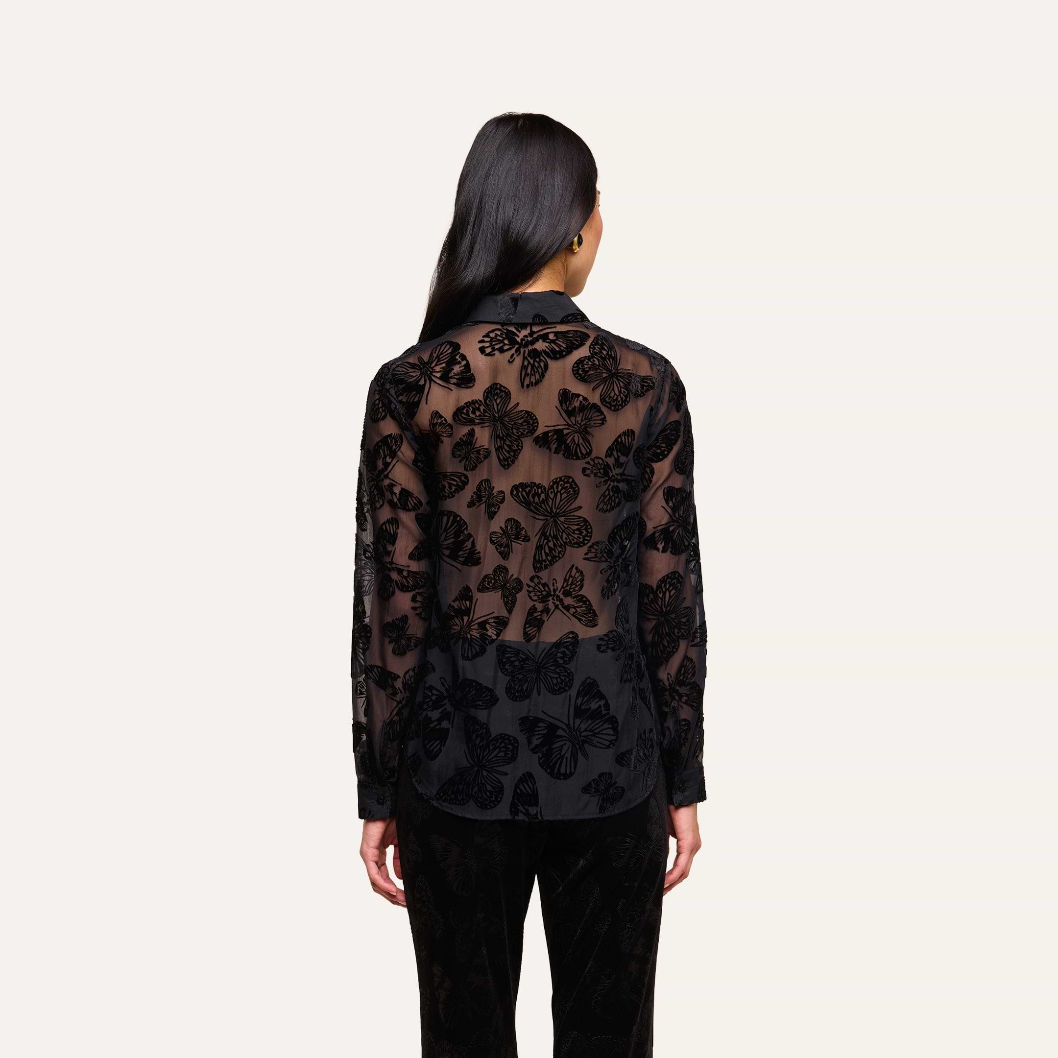 A woman with long dark hair faces away, wearing the Laurent Velvet Blouse—a relaxed fit button-down with a butterfly devoré pattern—and black pants, standing against a plain light background.