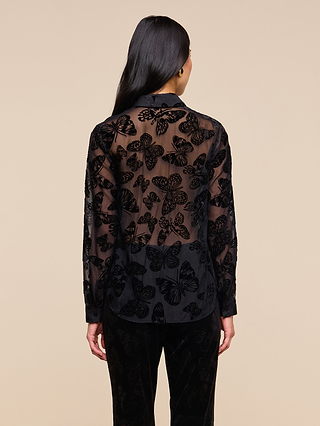 A person with long dark hair is seen from behind, standing against a plain beige background, wearing the Laurent Velvet Blouse—a sheer black burnout velvet top adorned with embroidered butterfly patterns.