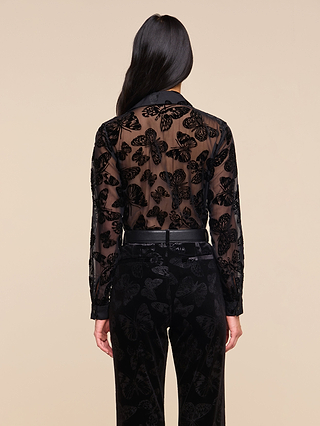 A person with long dark hair stands facing away, wearing the Laurent Velvet Blouse—a sheer black, relaxed fit top—paired with matching black pants featuring a velvet butterfly print on both pieces, set against a beige background.