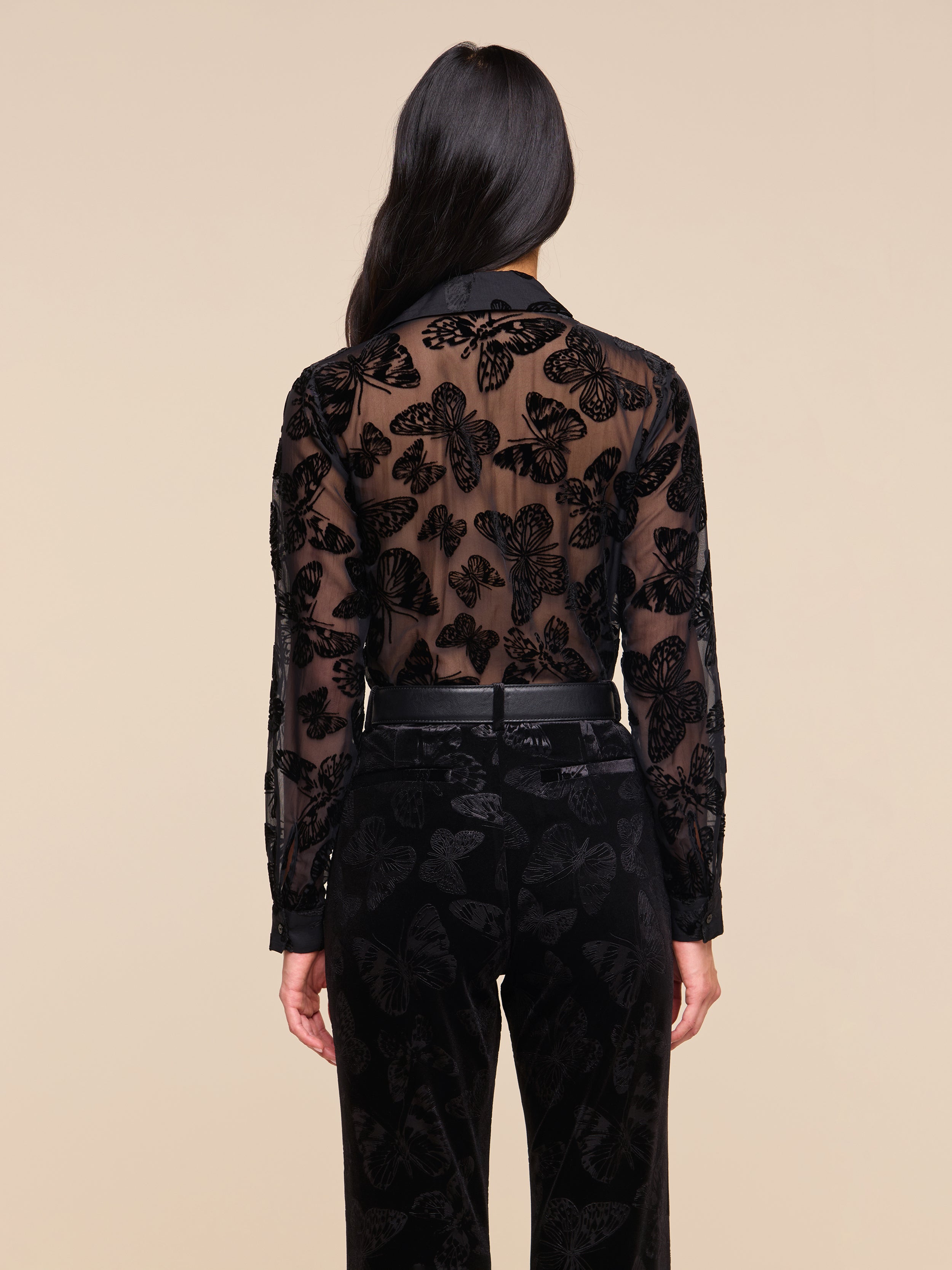 A person with long dark hair is shown from the back, wearing the Laurent Velvet Blouse featuring a butterfly pattern and black pants against a plain light background.