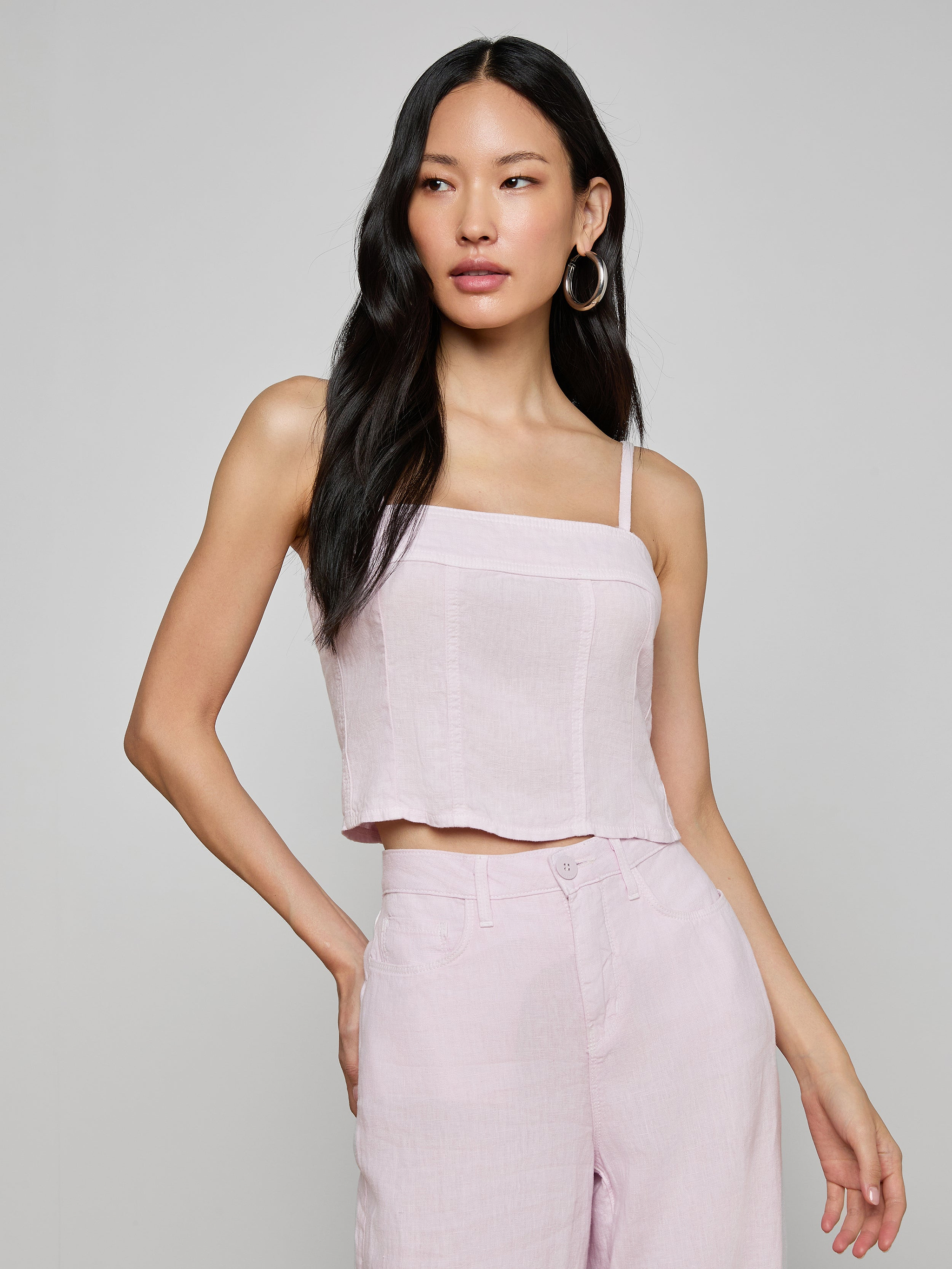 Wearing the Arlin Linen Tank, a woman with long black hair poses with one hand on her hip against a plain background, pairing the light pink cropped corset top with matching high-waisted pants.