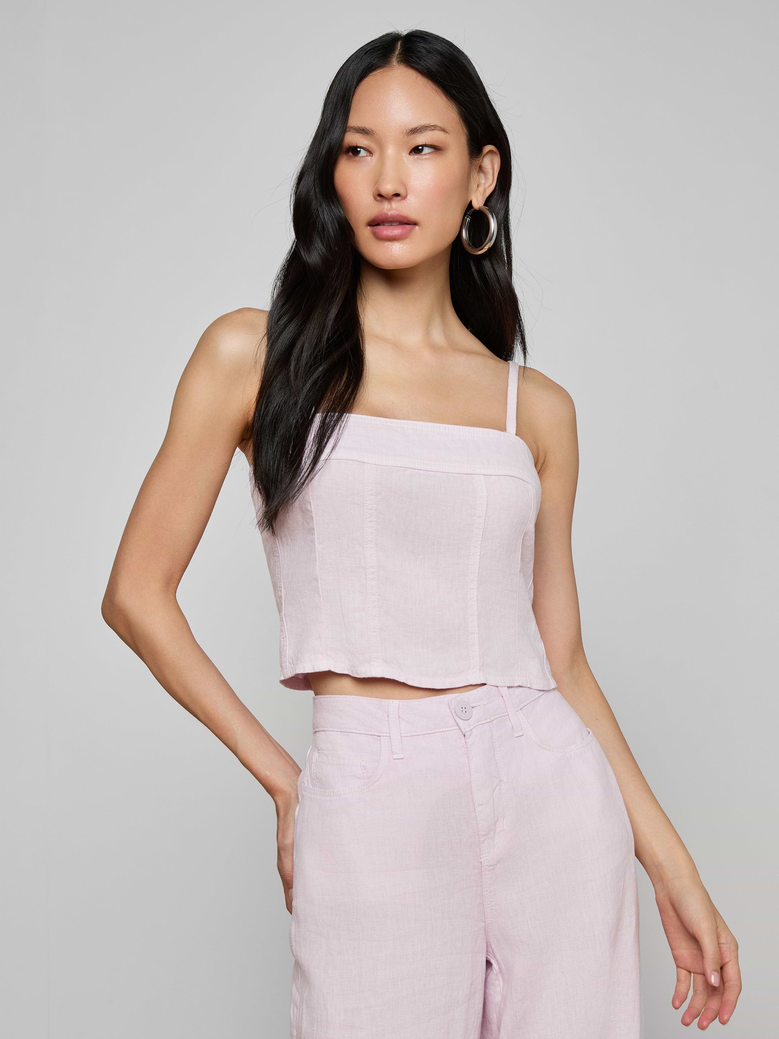 Wearing the Arlin Linen Tank, a woman with long black hair poses with one hand on her hip against a plain background, pairing the light pink cropped corset top with matching high-waisted pants.