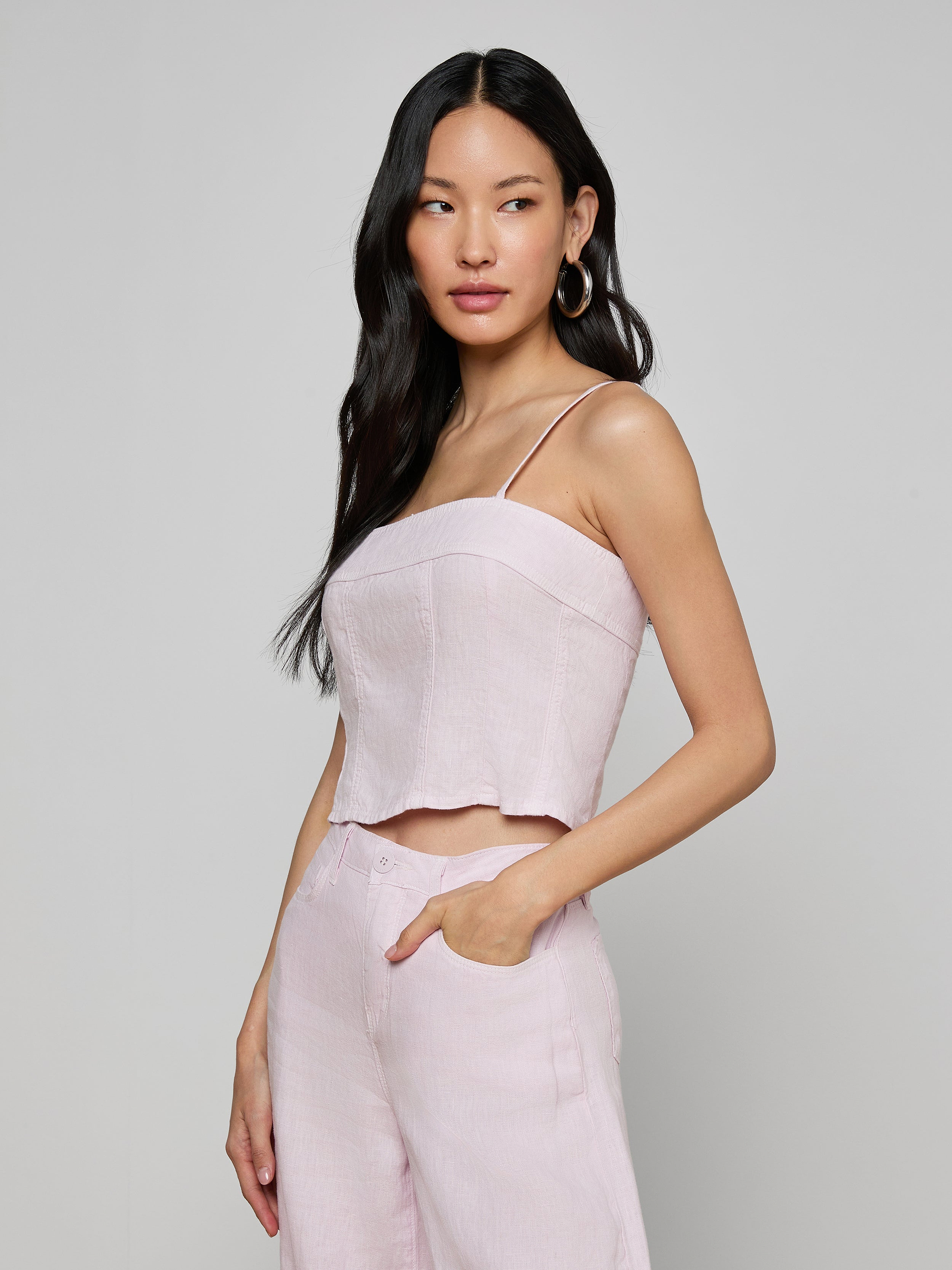 A woman with long dark hair wears the Arlin Linen Tank in light pink with matching pants, standing with one hand in her pocket against a plain, off-white background.