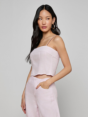 A woman with long black hair and gold hoop earrings models the Arlin Linen Tank, a light pink cropped top paired with matching high-waisted pants. She stands against a plain background, one hand in her pocket, looking to the side.