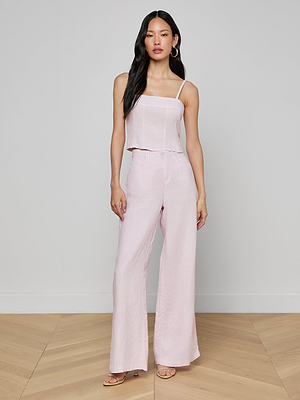 A woman stands indoors on a wooden floor wearing the Arlin Linen Tank, a light pink cropped corset-style top with spaghetti straps, paired with matching high-waisted wide-leg linen pants. She completes the look with gold hoop earrings and nude strappy heels.