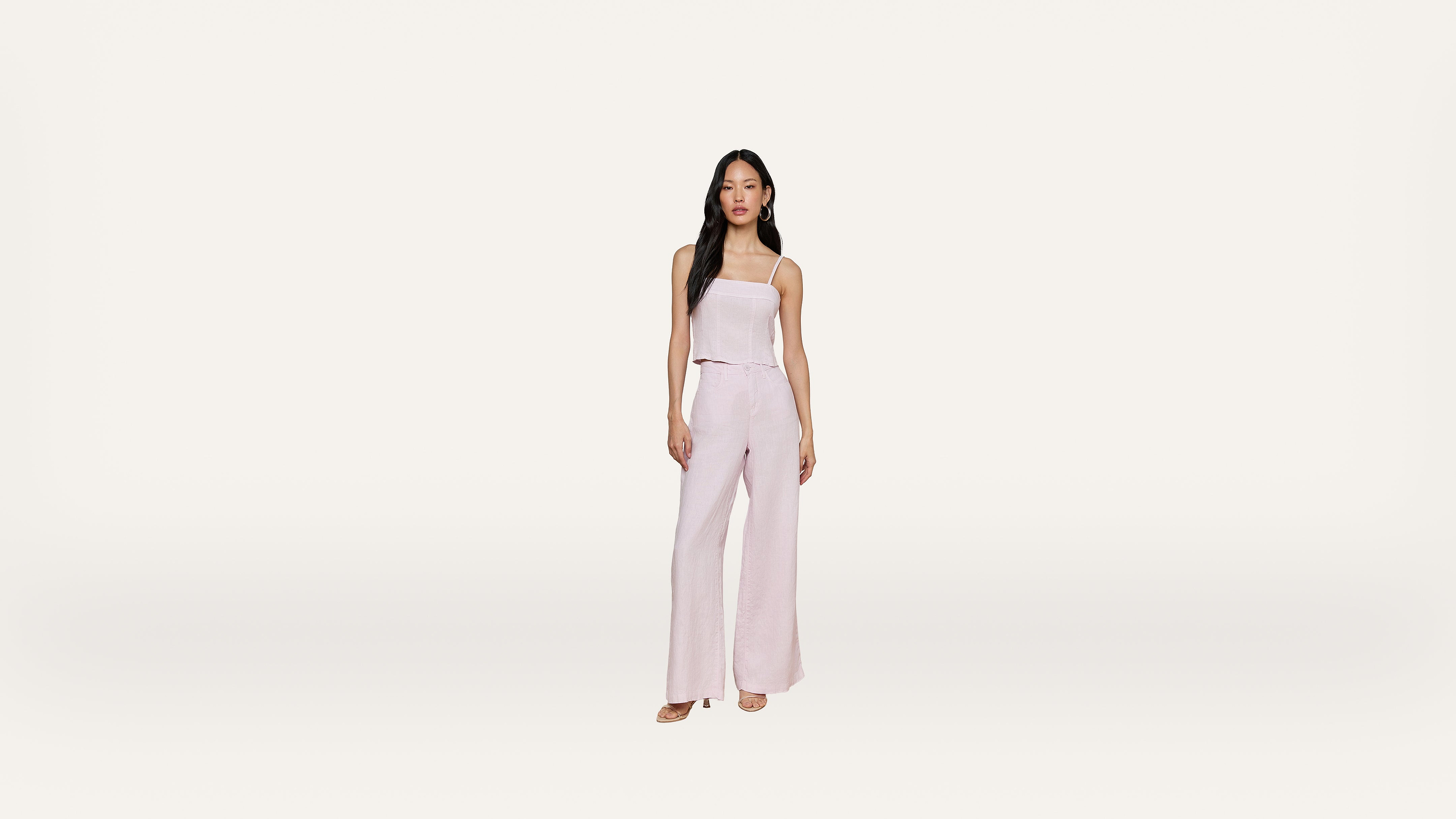 A woman stands against a plain white background, wearing a light pink, sleeveless top and matching wide-leg pants with open-toe shoes.
