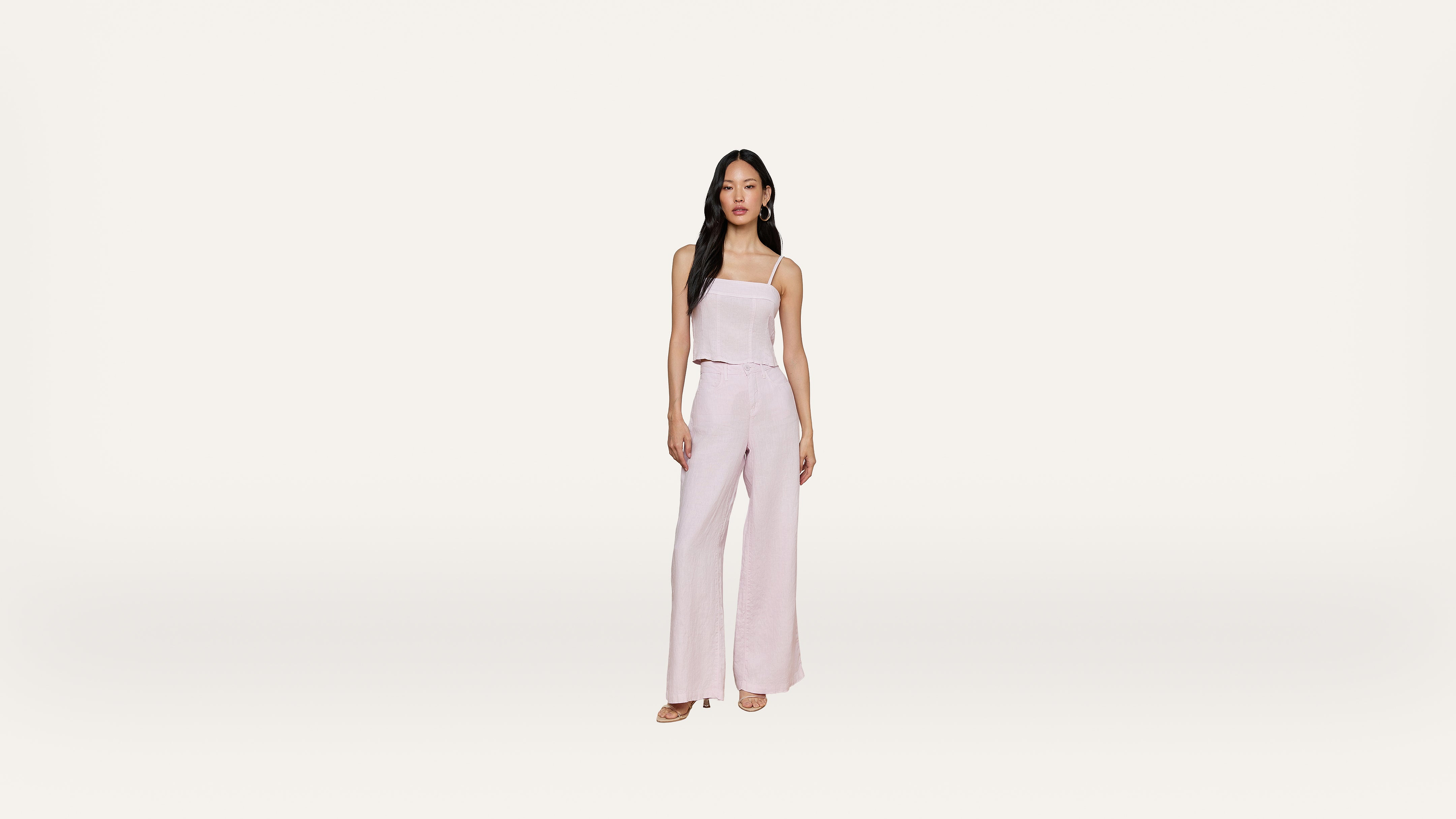 A woman stands against a plain white background, wearing a light pink, sleeveless top and matching wide-leg pants with open-toe shoes.