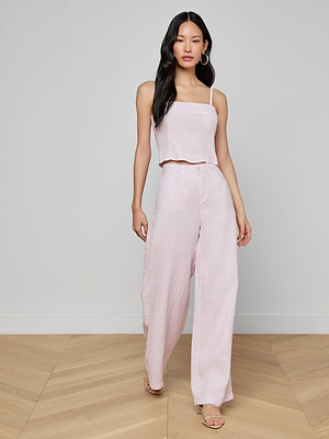 A woman with long dark hair and large hoop earrings stands on a wooden floor against a light background, wearing the Arlin Linen Tank in light pink paired with matching high-waisted wide-leg pants and heeled sandals.