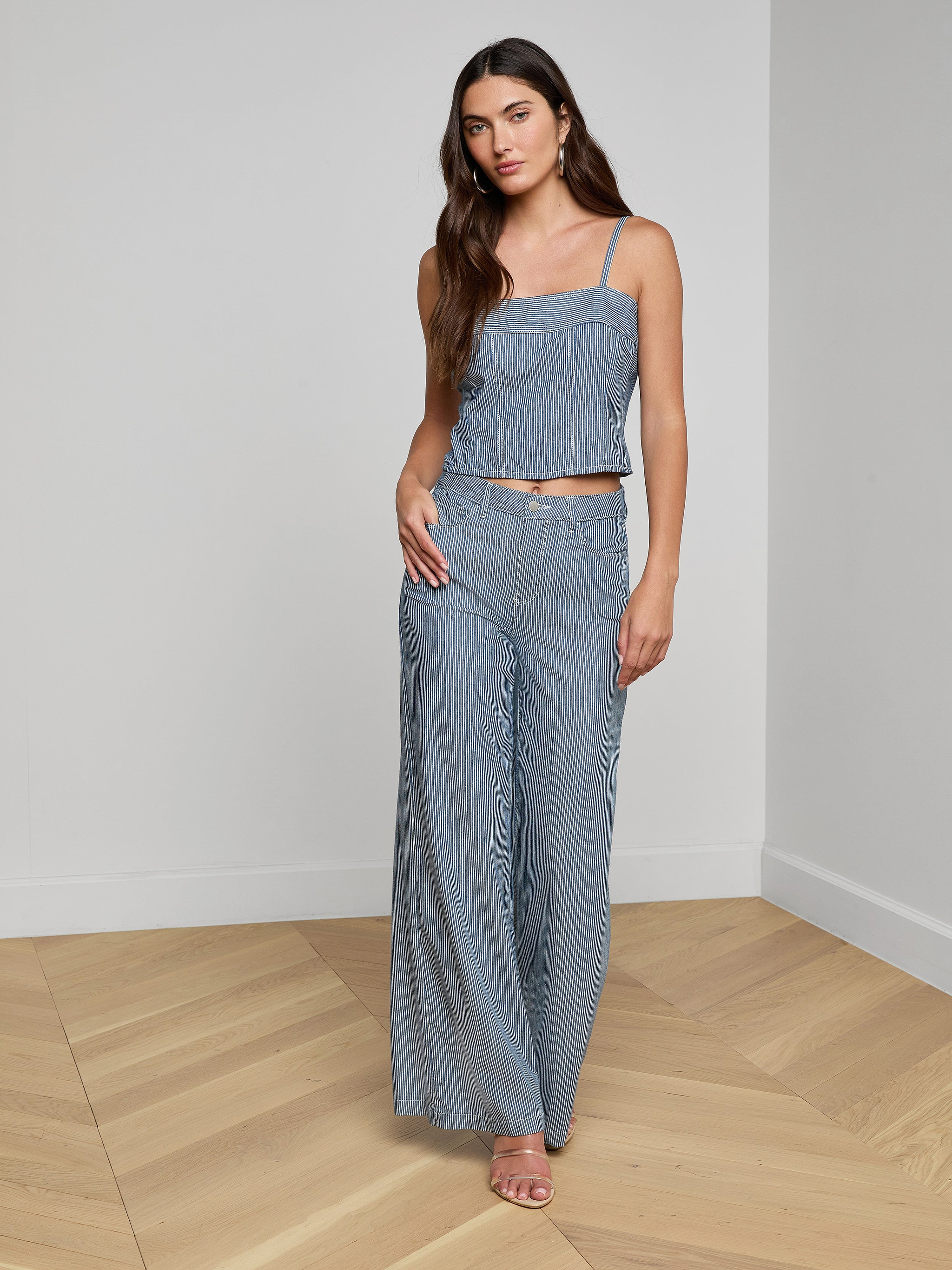 A woman wearing the Arlin Tank— a sleeveless denim corset top— paired with matching wide-leg pants, stands against a plain white background with one hand on her hip and the other relaxed by her side.