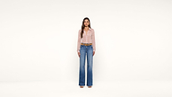 A woman stands against a plain white background, wearing a light pink blouse, wide-leg blue jeans, brown belt, and beige open-toe shoes. She has long brown hair and faces the camera directly.