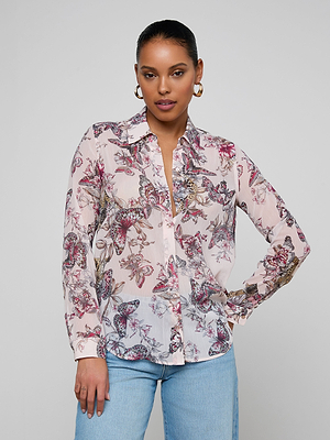 A woman wears gold hoop earrings, light blue jeans, and the Laurent Blouse—a sheer poly chiffon, relaxed-fit, long-sleeve button-up shirt with a pink and purple floral print—against a plain light background.