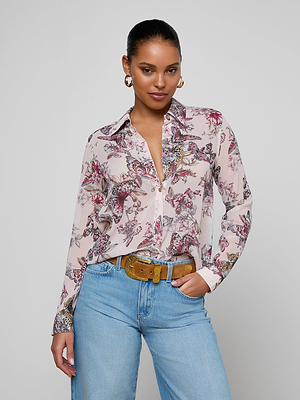 A woman stands confidently against a plain background, wearing the Laurent Blouse—a sheer poly chiffon button-down with a butterfly jungle print in pink and burgundy accents—paired with light blue jeans, a tan belt, and hoop earrings. Her hair is pulled back.