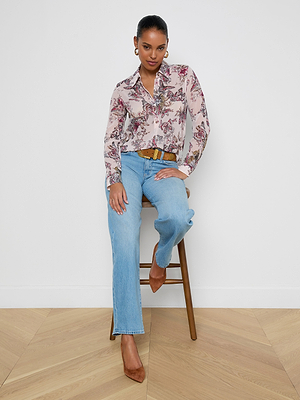 A woman with her hair pulled back and a slight smile wears the Laurent Blouse—a relaxed fit, long sleeve, light pink floral top—paired with light blue jeans, a brown belt, and brown heels while sitting on a wooden stool against a white wall.