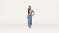 A woman stands against a plain white background, wearing a sleeveless denim jumpsuit and sandals. She has long dark hair and her left hand is in her pocket.