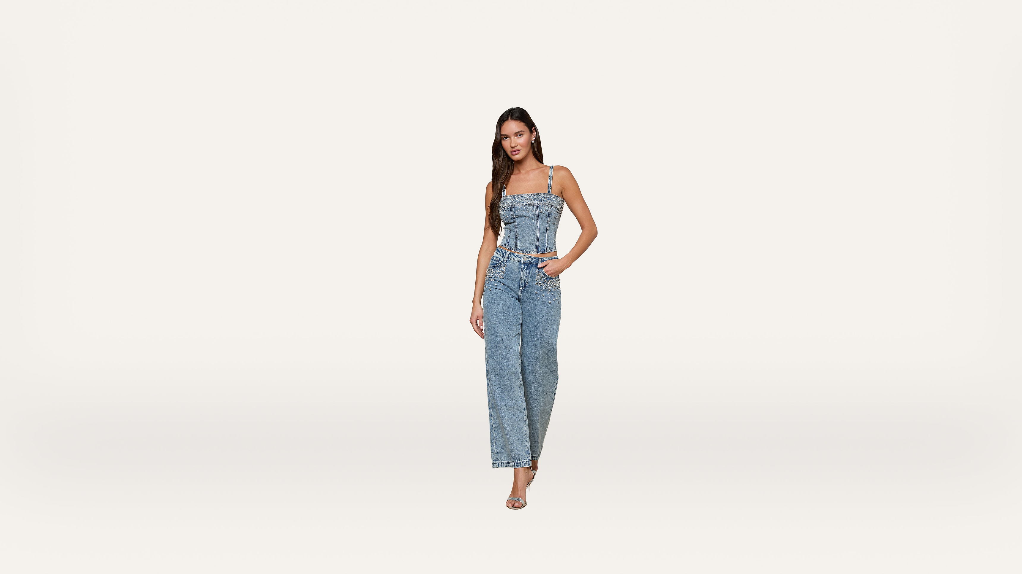 A woman stands against a plain white background, wearing a sleeveless denim jumpsuit and sandals. She has long dark hair and her left hand is in her pocket.