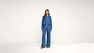A woman stands in a studio wearing a blue button-up shirt and matching wide-leg pants with a black belt and sandals against a plain white background.