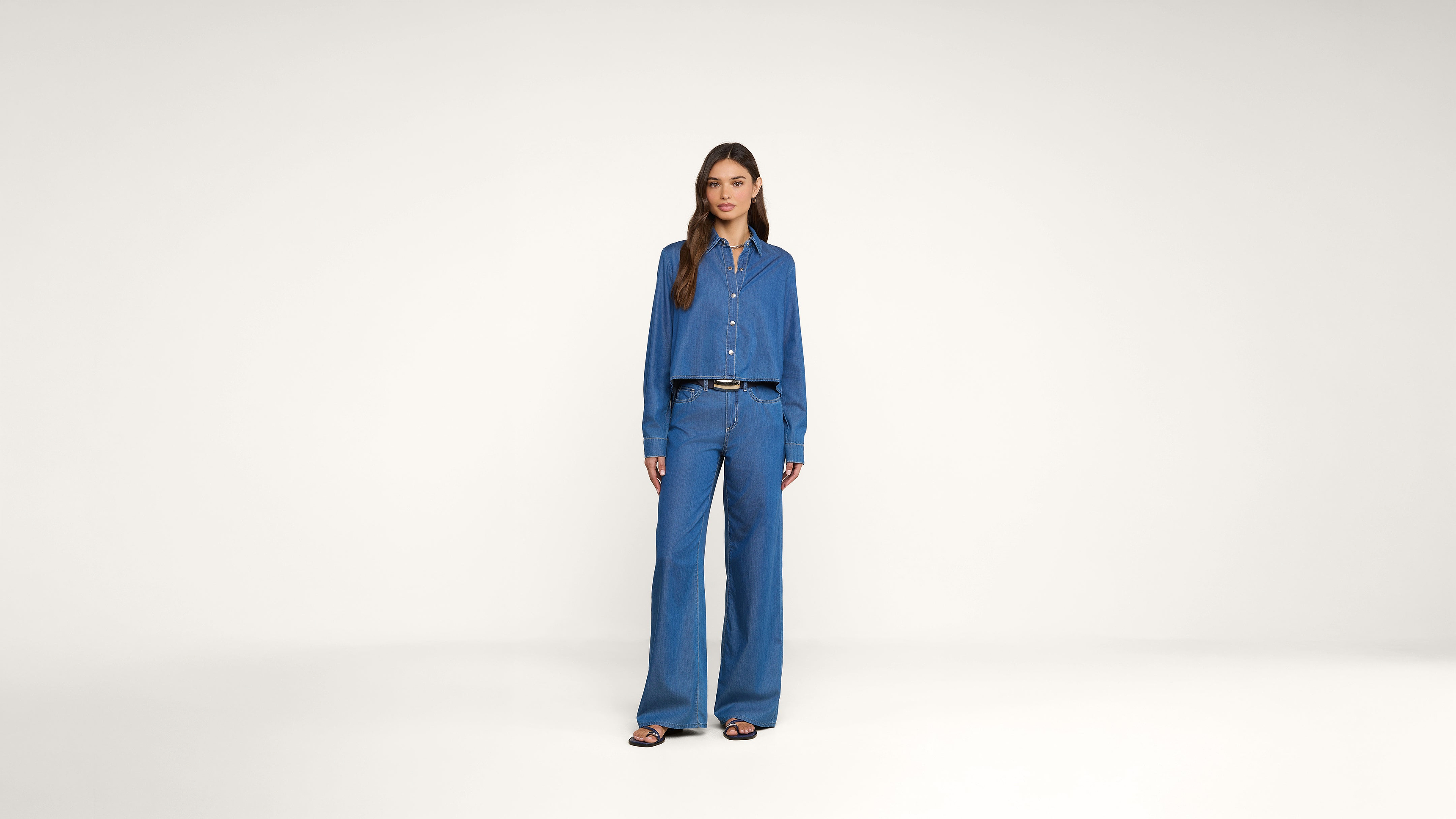 A woman stands in a studio wearing a blue button-up shirt and matching wide-leg pants with a black belt and sandals against a plain white background.