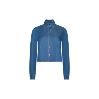 The Annette Chambray Shirt is shown on a plain white background. This blue denim long-sleeve button-up shirt features a pointed collar, front buttons, and a chest pocket. Designed with a relaxed fit and a subtle high-low hem, it offers classic chambray style and everyday comfort.