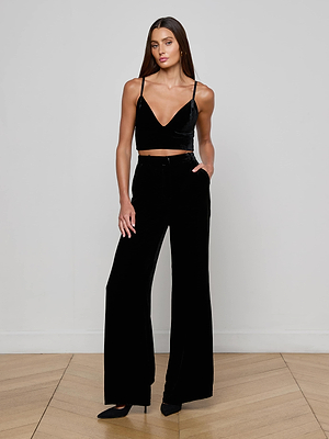 A woman with long brown hair wears the L'AGENCE Aislin Velvet Bralette, black high-waisted wide-leg pants, and black pointed-toe shoes, posing against a plain white wall with one hand in her pocket.