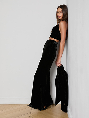 A woman with long brown hair wears the L'AGENCE Aislin Velvet Bralette in black, paired with high-waisted velvet wide-leg pants and a matching jacket, standing against a white wall in black high heels.