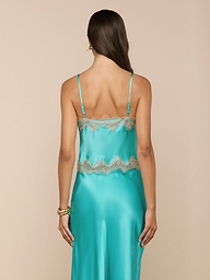 A woman with long brown hair faces away, wearing the Kimber Silk Lace Camisole—a turquoise satin piece featuring thin straps and delicate beige lace trim. She stands against a plain light background.
