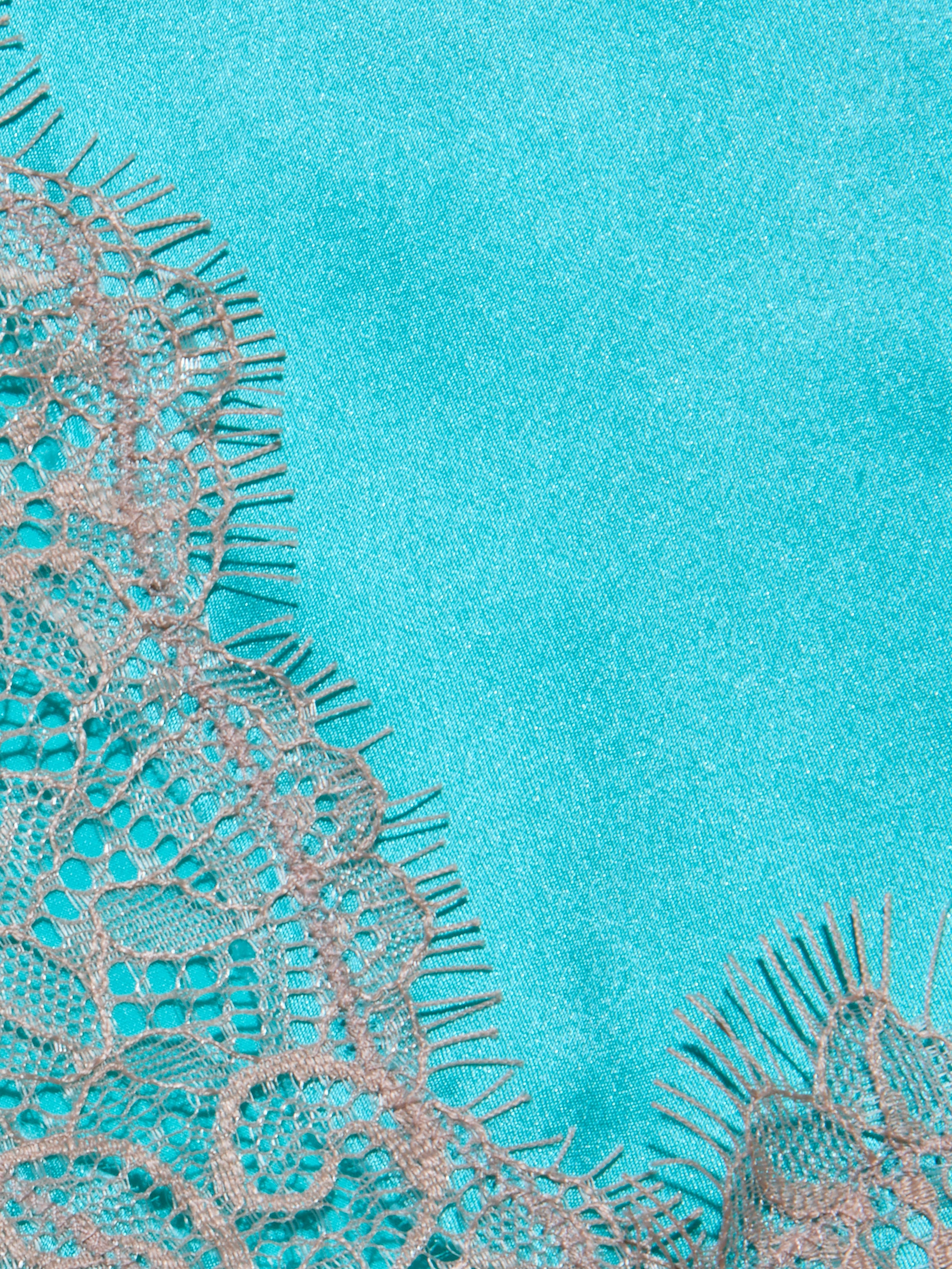 Close-up of the Kimber Silk Lace Camisole, showcasing delicate beige lace with intricate floral patterns against a smooth, bright turquoise background. The camisole’s frayed edges create a soft contrast with the vibrant, solid-colored fabric beneath.