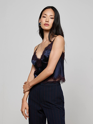 A woman with long black hair models the L'AGENCE Kimber Silk Lace Camisole in navy, paired with dark pinstriped trousers, standing against a light gray background and gazing confidently at the camera with one arm across her waist.