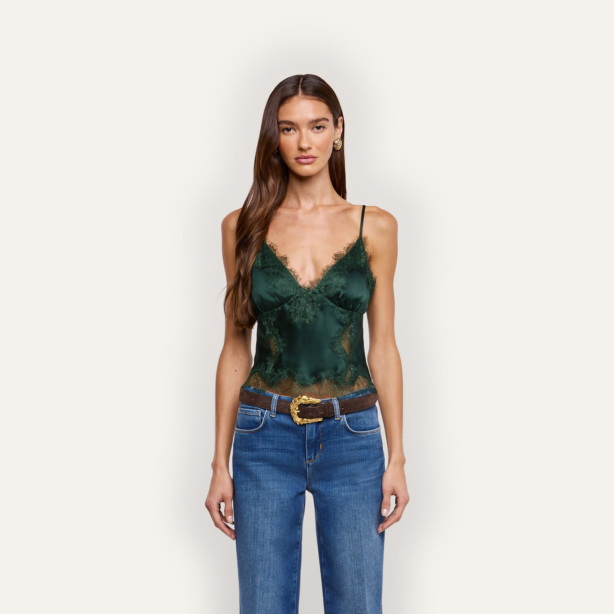 A woman wears a dark green Kimber Silk Lace Camisole with a deep V-neckline and lace trim, tucked into blue jeans with a brown leather belt.