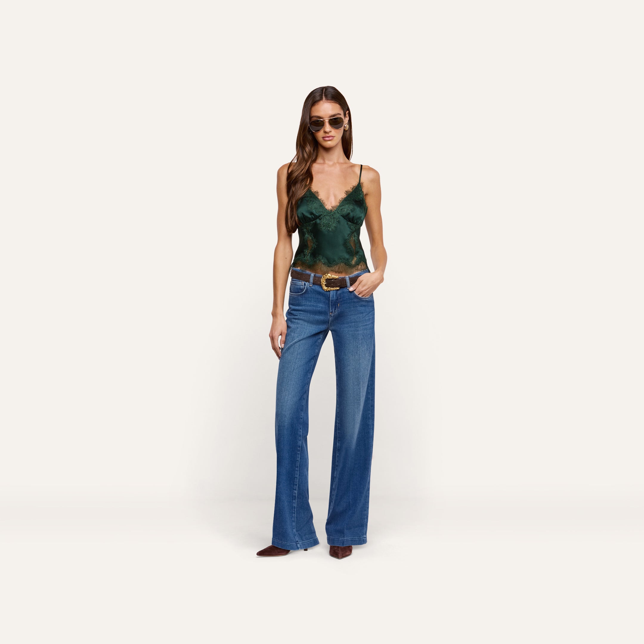 A person wearing a dark green lace-trimmed Kimber Silk Lace Camisole with a deep V-neckline, wide-leg blue jeans with a brown belt, and sunglasses.