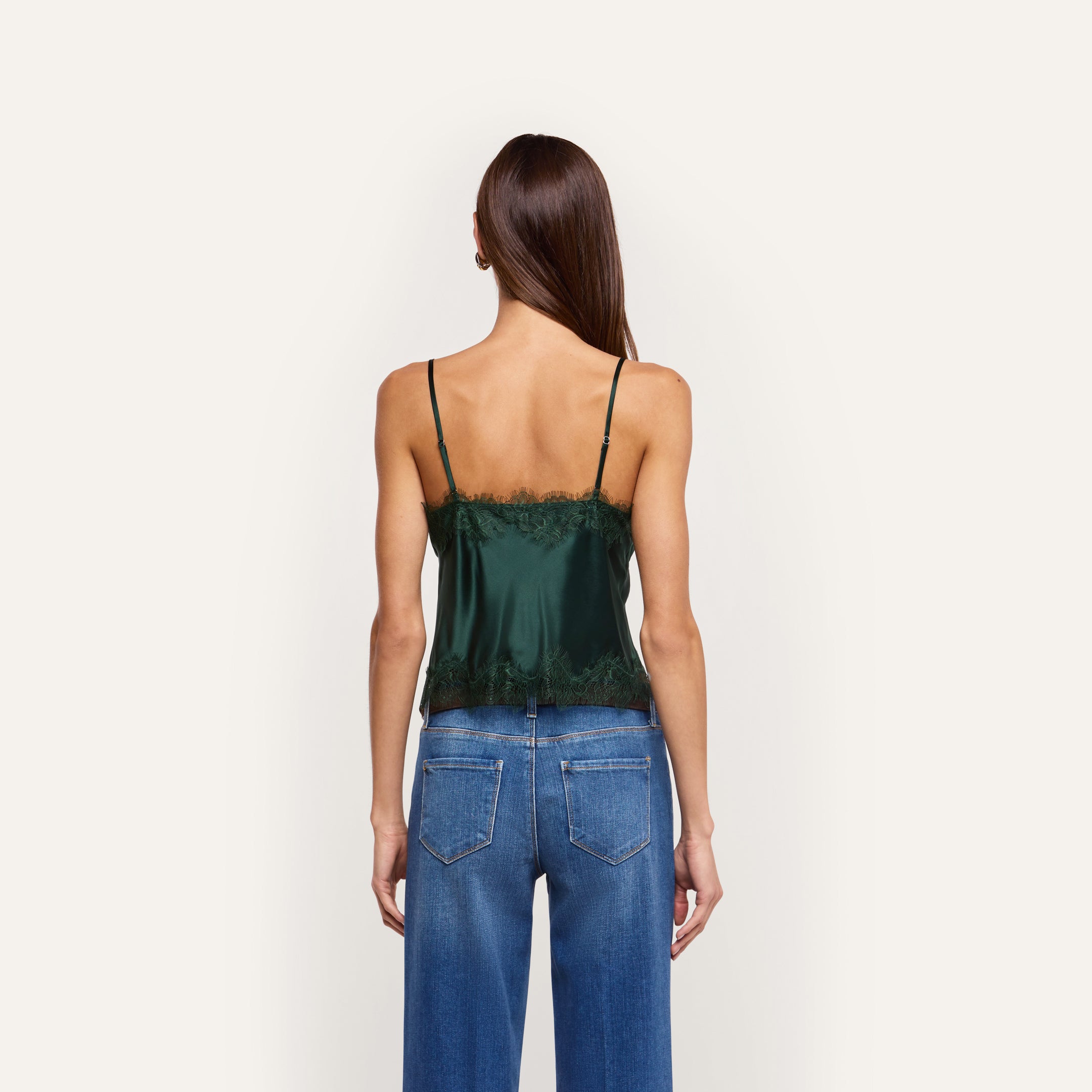 Woman viewed from behind wearing a dark green silk Kimber Silk Lace Camisole with black lace trim and blue jeans, featuring a deep V-neckline.