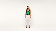 A woman stands against a plain white background, wearing a green lace camisole top, white high-waisted pants, a black belt, and black heels. She faces forward with arms relaxed by her sides.
