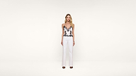 A woman stands facing forward against a plain white background, wearing a white top with black lace details, white high-waisted pants, and black open-toe heels.
