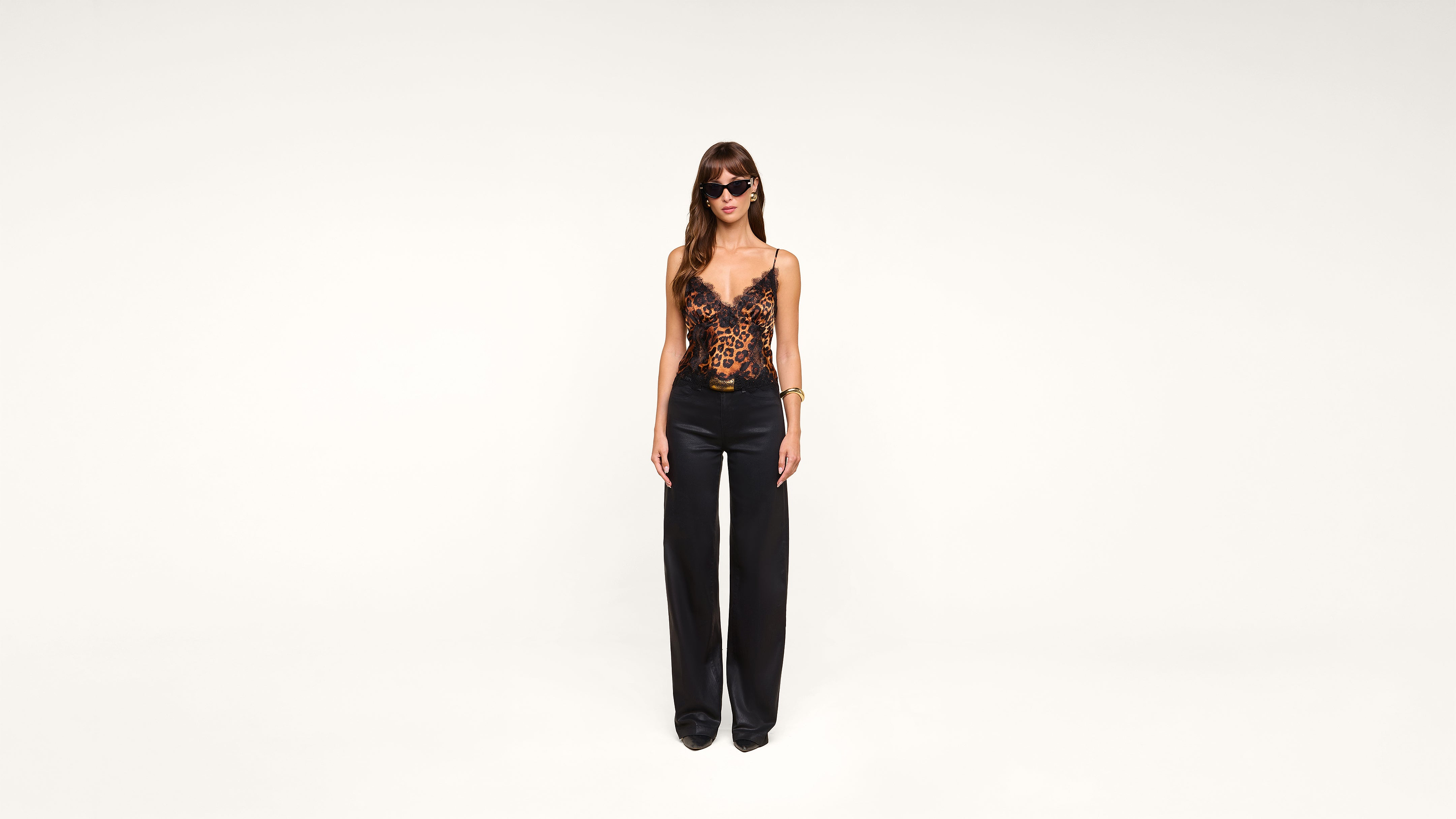 A woman stands against a plain white background, wearing sunglasses, a black and orange lace camisole top, black wide-leg pants, and black shoes.