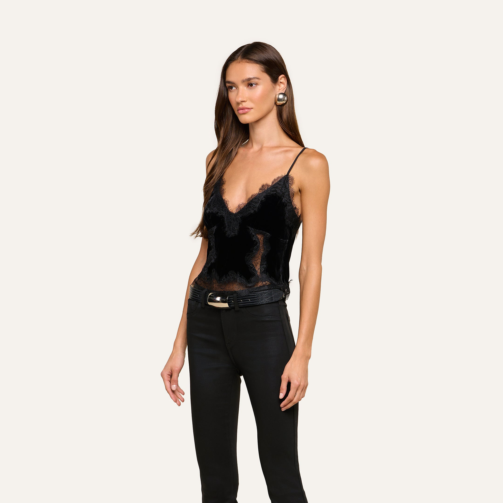 A woman with long brown hair is wearing the Kimber Velvet Lace Camisole, paired with black pants featuring a belt and large earrings, standing against a plain light background.