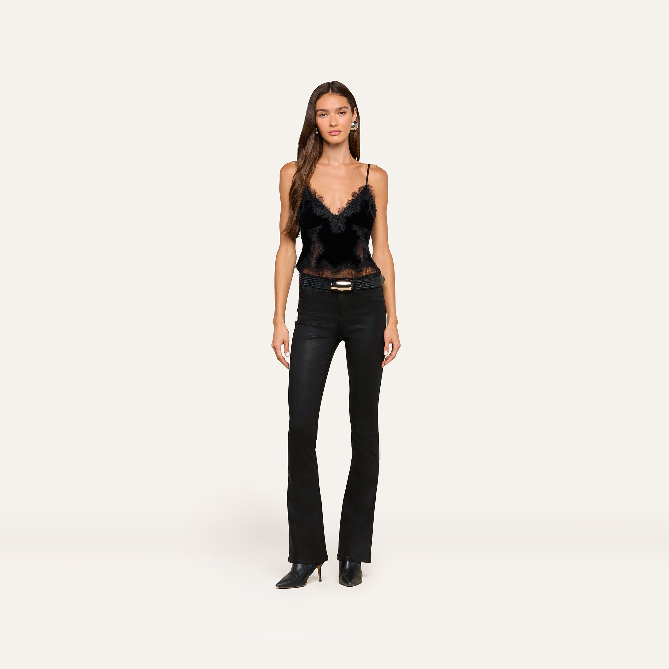 A woman with long brown hair and a neutral expression stands against a plain background, wearing the Kimber Velvet Lace Camisole, paired with black fitted jeans, a belt, and black pointed-toe boots.