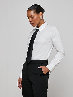 A woman with short, slicked-back hair stands sideways against a light gray background, wearing the white Blake Bodysuit and black pants with a removable tie, one hand in her pocket.