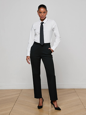 A woman stands confidently in the Blake Bodysuit, styled with a removable tie and black pants, black heels on a light wood floor against a white wall, one hand casually in her pocket.