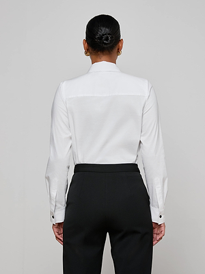 A person with dark hair in a neat bun is shown from behind, wearing the Blake Bodysuit—a white organic cotton bodysuit with snap closures—paired with black pants against a plain, light-colored background.