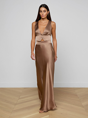 A woman stands on a wooden floor against a plain white wall, wearing the Devony Silk Vest—a sleeveless, deep V-neck bronze satin piece with a fitted waist and floor-length skirt—evoking elegant evening style.