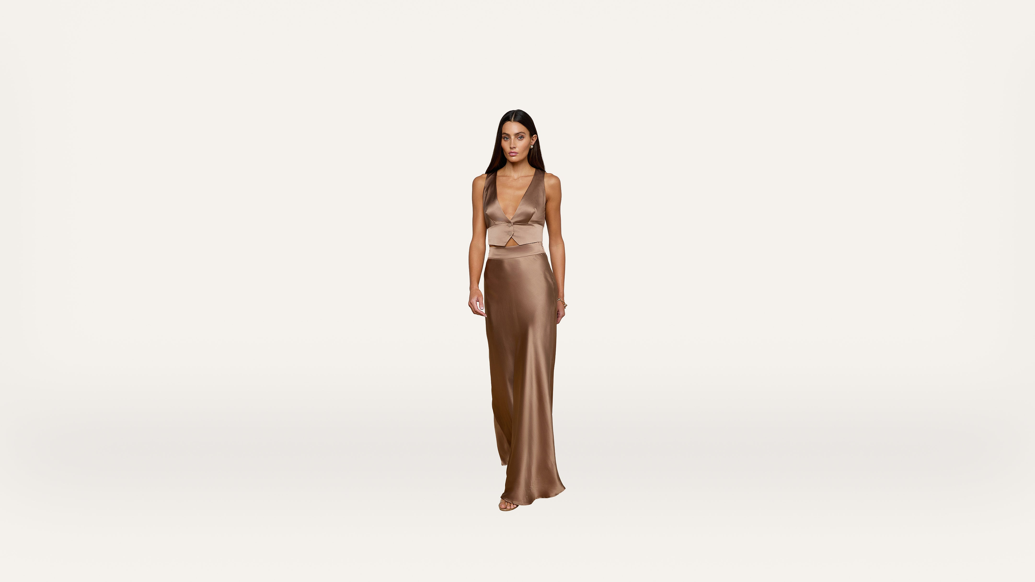 A woman with long dark hair wears a sleeveless, light brown silk crop top and a matching long skirt, standing against a plain white background.