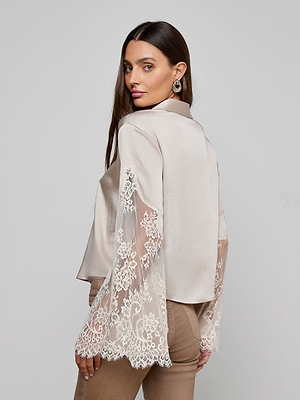 Woman with long brown hair wears the Capri Lace Sleeve Blouse—a silky beige bohemian top with sheer lace bell sleeves—paired with tan pants, standing against a plain background and looking over her shoulder at the camera.