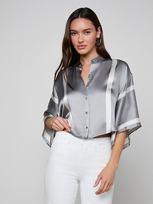 A woman with long brown hair wears the Dru Silk Blouse, featuring kimono sleeves and white stripes, paired with high-waisted white pants. She poses against a plain light gray background with one hand on her hip.